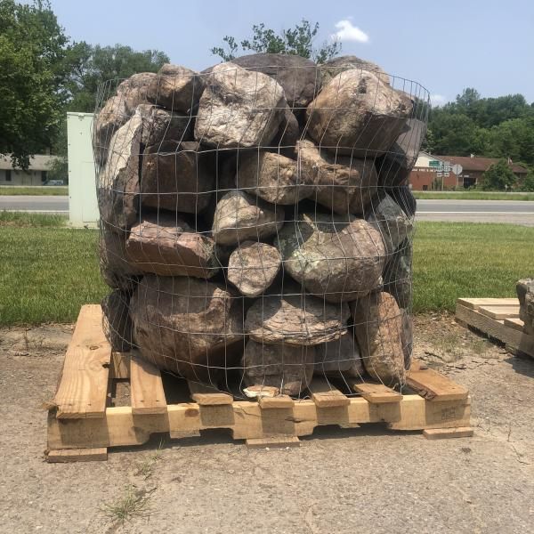 Stacked landscaping rocks in a wire cage on a wooden pallet outdoors beside a road