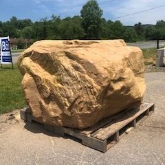 Large tan boulder on a wooden pallet in a parking lot, with trees and a roadside sign in the background