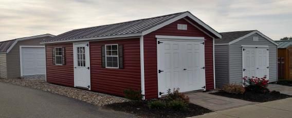 Small red-and-white shed with white garage doors beside gray sheds in a paved lot