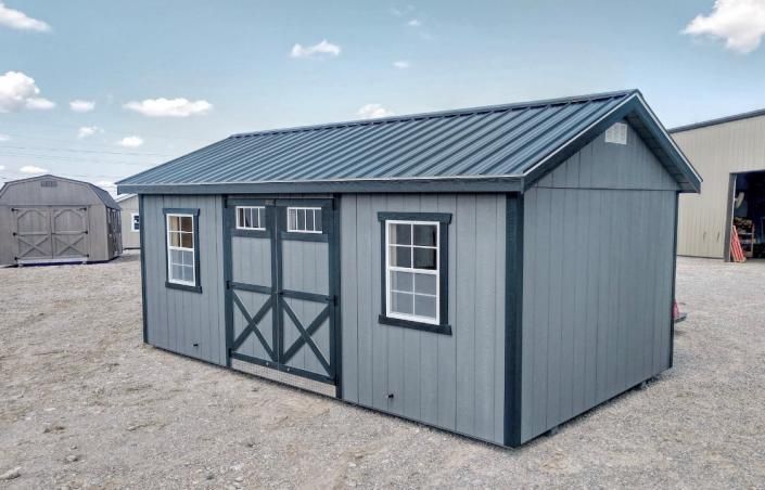 Small gray shed with white-trimmed windows and double doors on a gravel lot