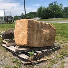 Large tan boulder on wooden pallets beside a roadside grassy area under a blue sky