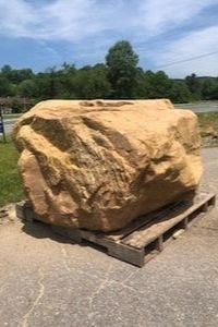 Large tan boulder resting on a wooden pallet outdoors on a paved surface under a partly cloudy sky