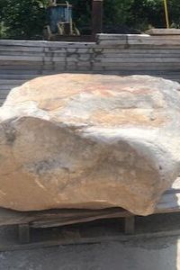 Large pale boulder resting on a wooden pallet outdoors, with stacked stone wall in the background