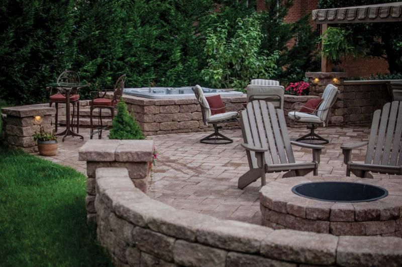 Stone patio with outdoor fire pit, cushioned chairs, and potted plants surrounded by greenery.