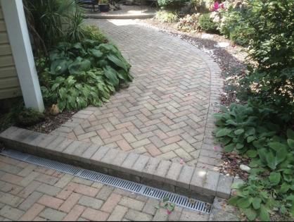 Curved brick garden path bordered by lush greenery and a drainage grate