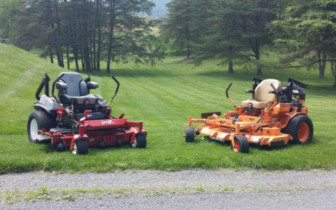 Two riding lawn mowers, one red and one orange, parked on grass near a gravel path.