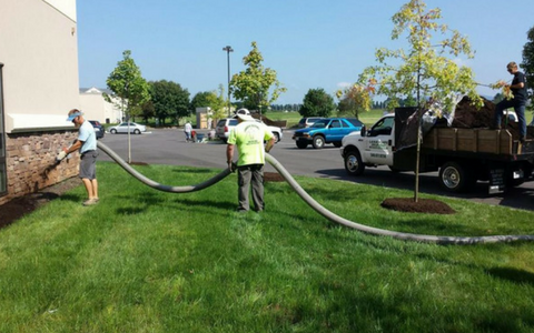 Workers in safety gear lay hoses across a grassy lot beside parked trucks and a building.