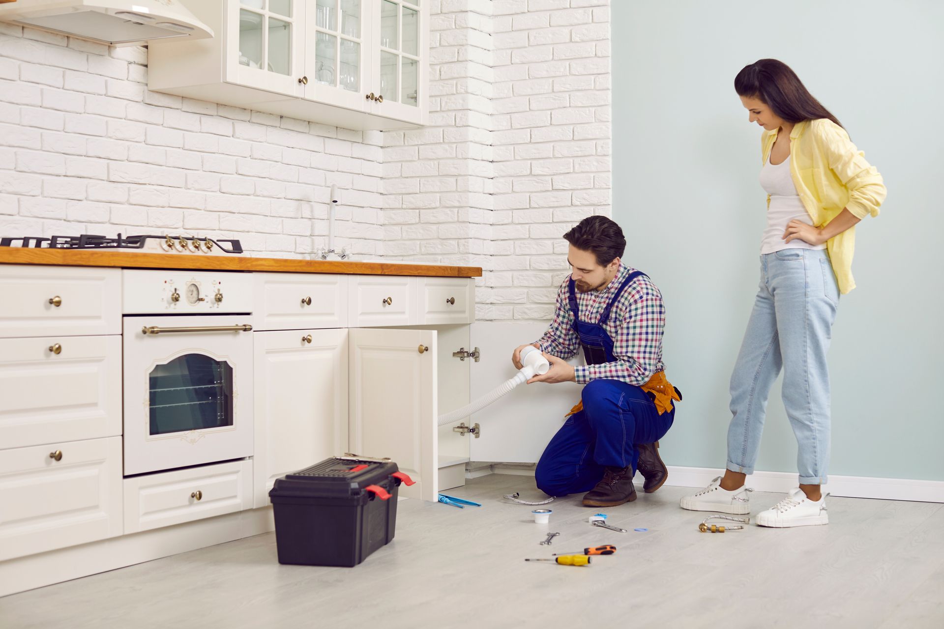 A plumber kneels under a kitchen cabinet, repairing pipes, while the homeowner stands nearby