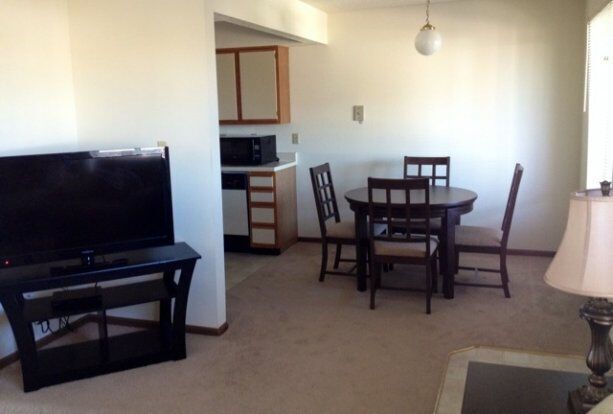 Living room with kitchen in background; TV, dining table, beige carpet, white walls.