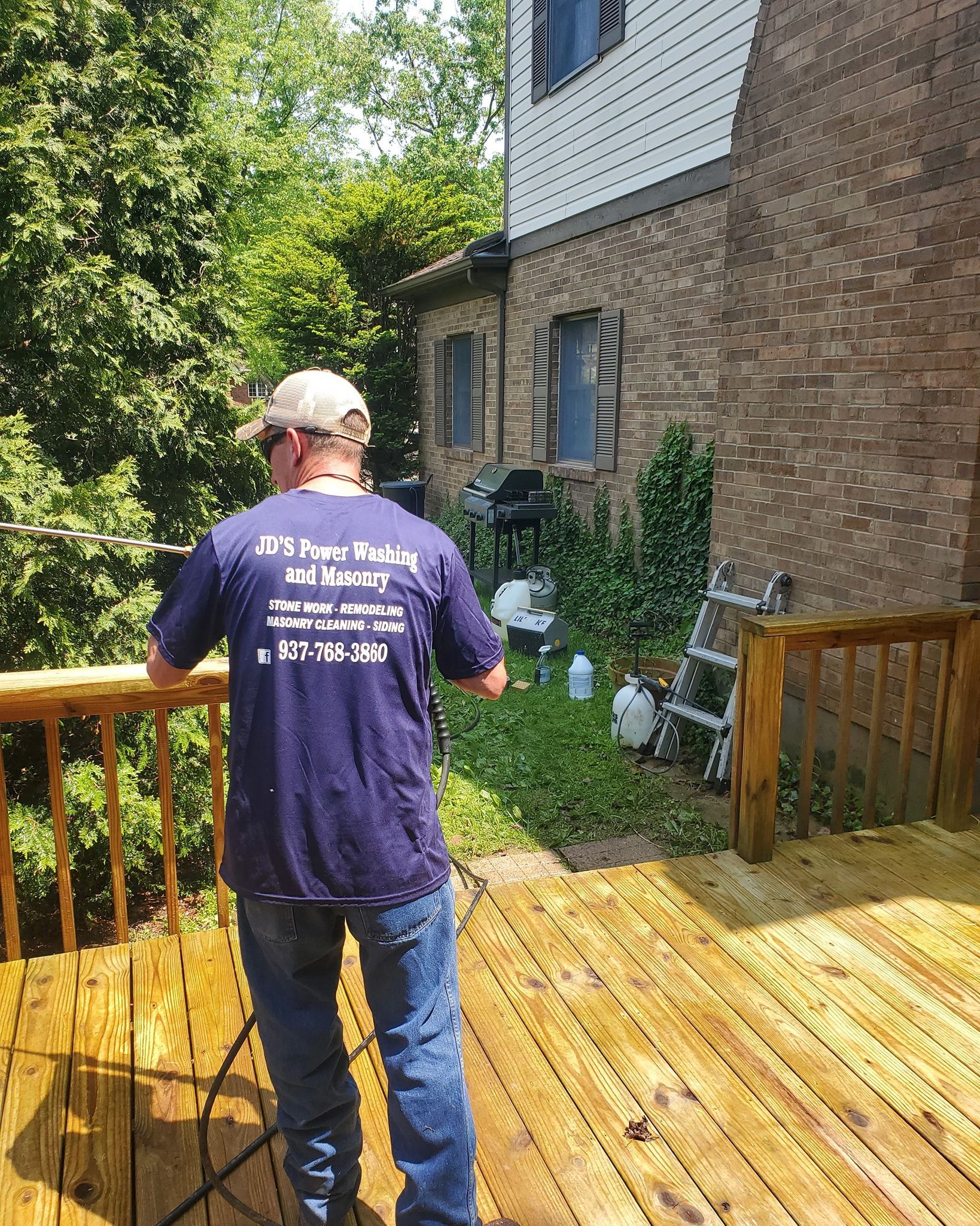 A person in a blue t-shirt and jeans stands on a wooden deck overlooking a wooded area next to a brick house.