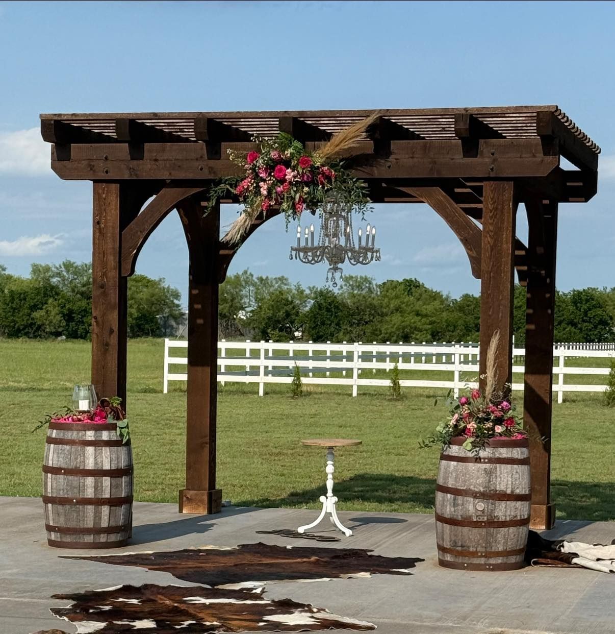 Rustic wooden wedding arbor with floral decorations, chandelier, and barrels in a grassy field.