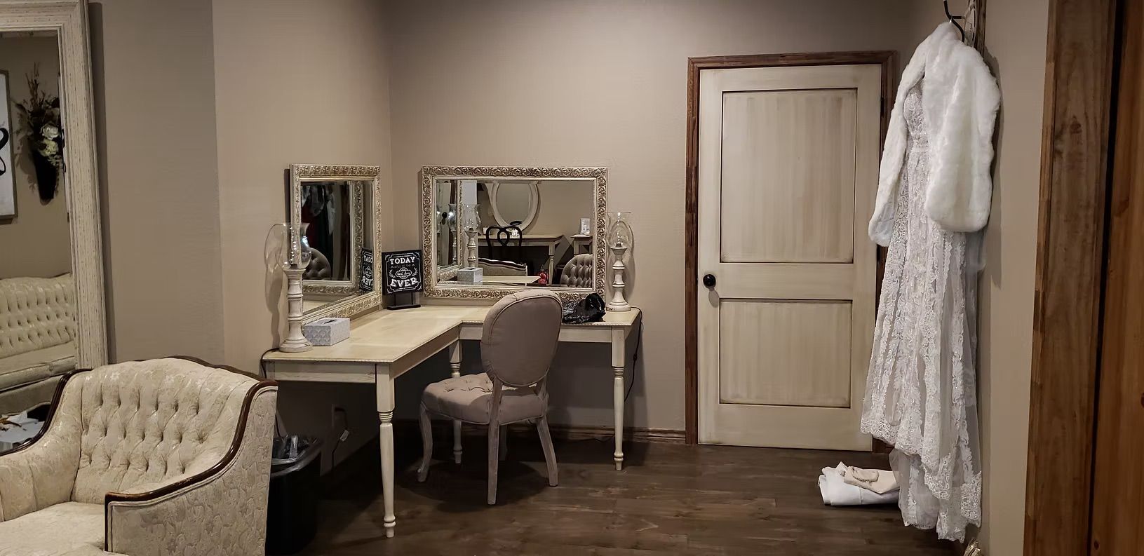 A vanity with mirrors, a chair, and a white dress hanging near a door.