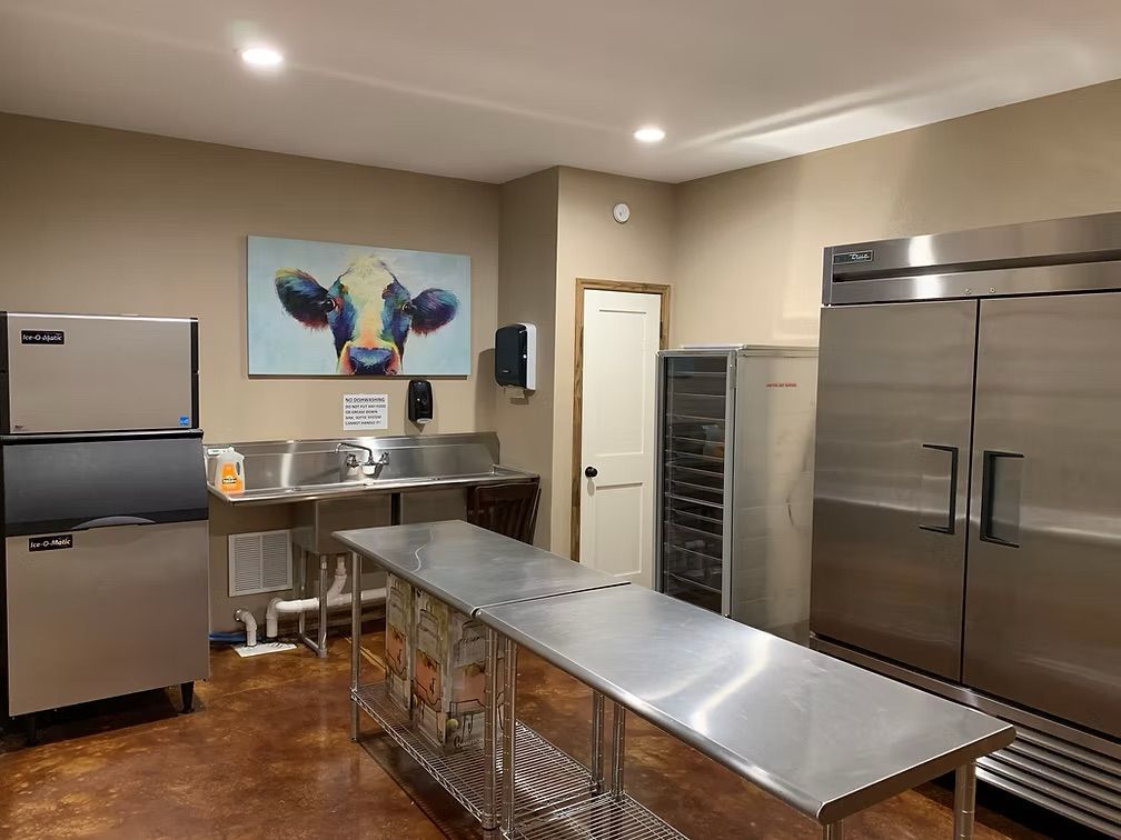 Commercial kitchen with stainless steel appliances, prep table, sinks, cow painting, and door.