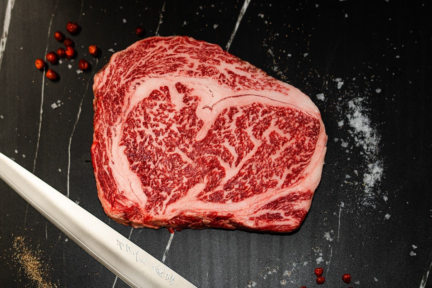 Raw marbled steak on a dark surface with scattered salt and red peppercorns