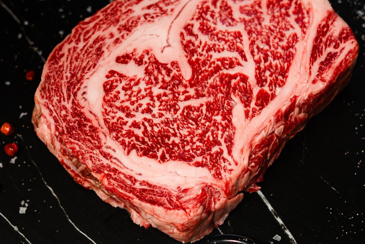 Raw marbled ribeye steak on a dark surface, with red seasoning flecks