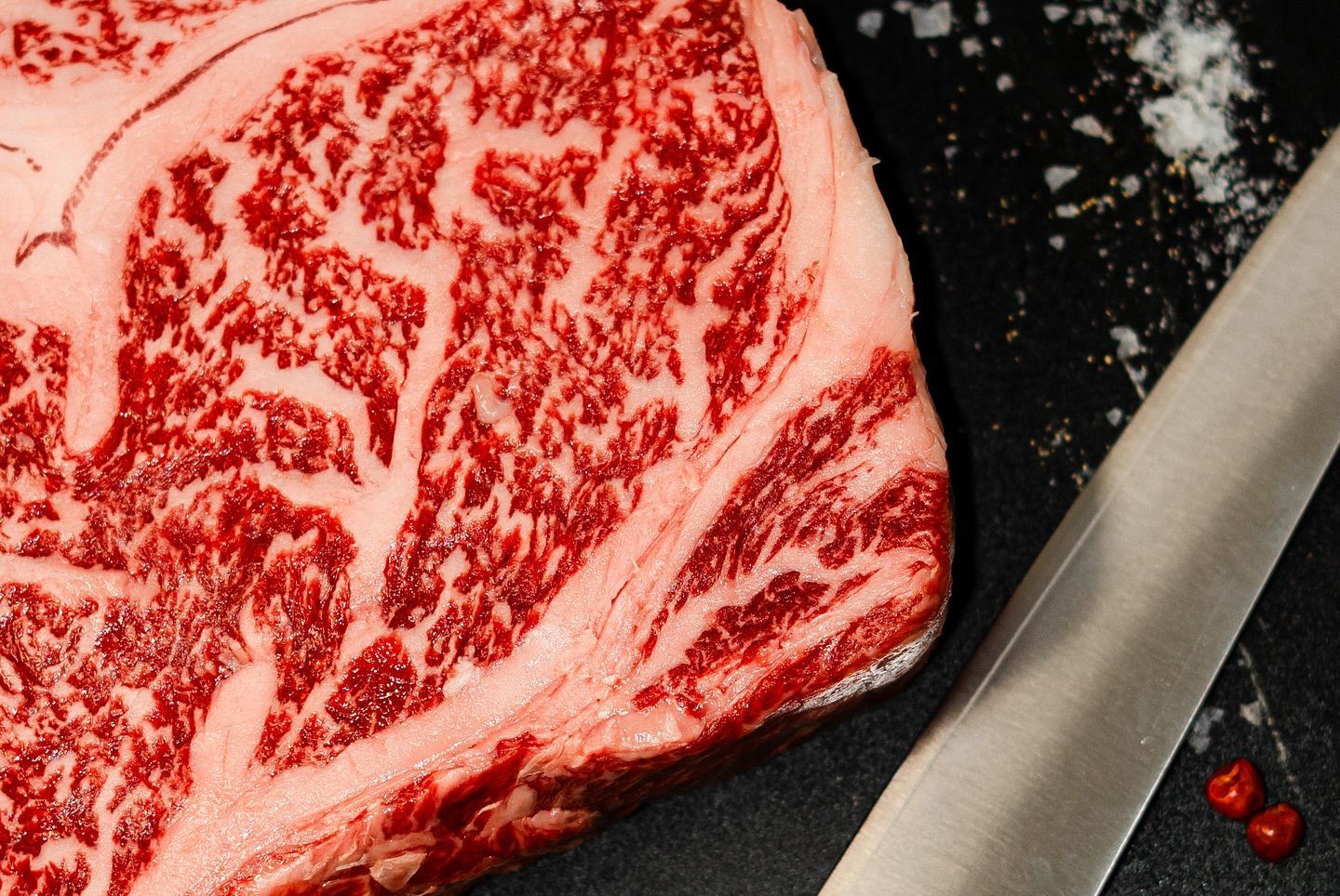 Raw marbled steak on a black surface beside a knife and peppercorns