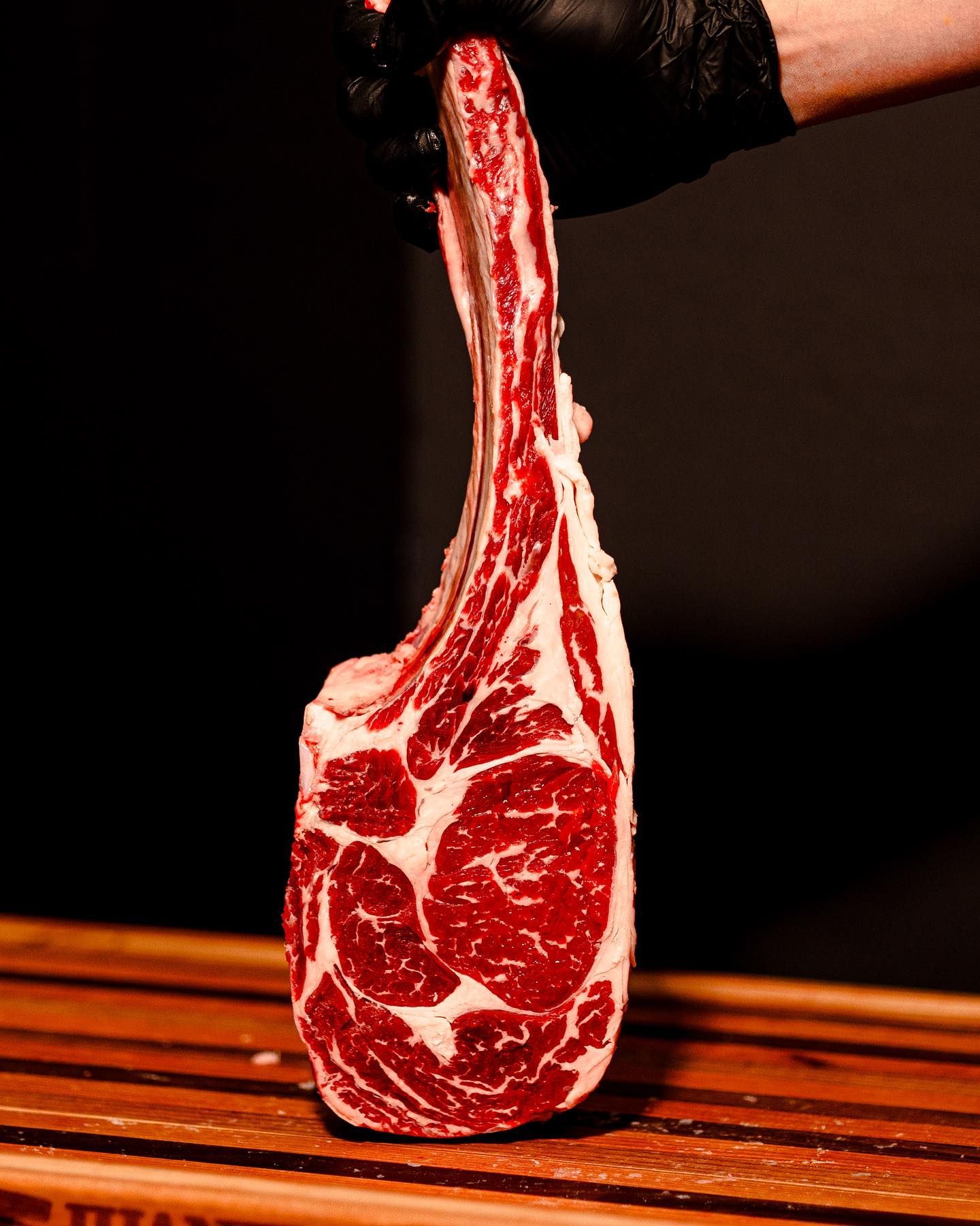 Gloved hand holding a raw ribeye steak above a wooden cutting board