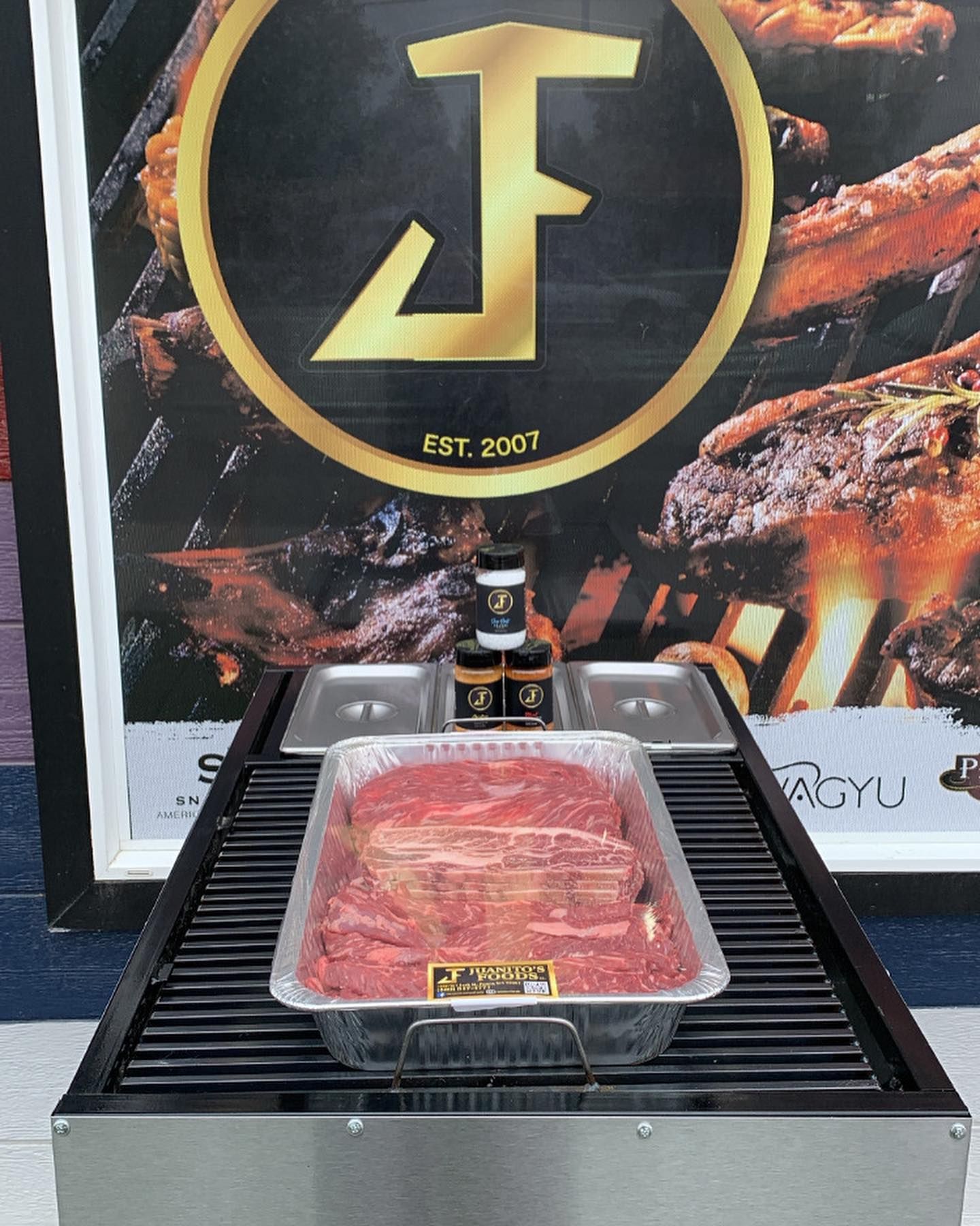 Fresh steak tray on a black grill display with a gold logo backdrop and sauce bottle.