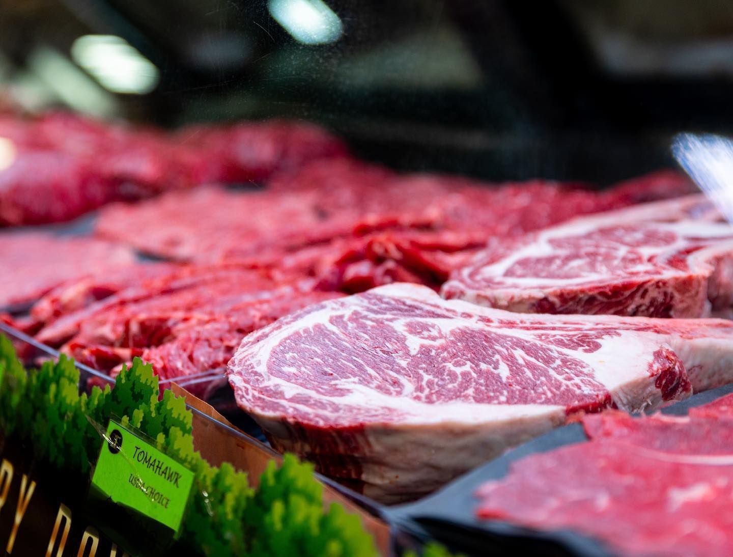 Butcher case with assorted raw red steaks and ribeye cuts on display, with green garnish and price tags