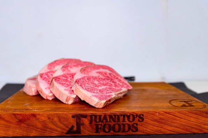 Raw marbled steaks on a wooden cutting board with “Juantos Foods” logo