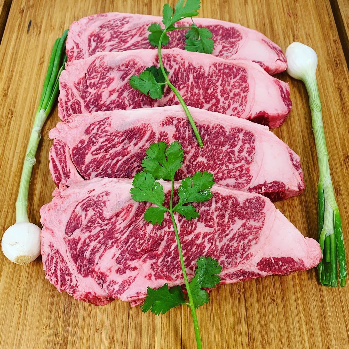 Four marbled raw steaks on a wooden board, garnished with cilantro and green onions