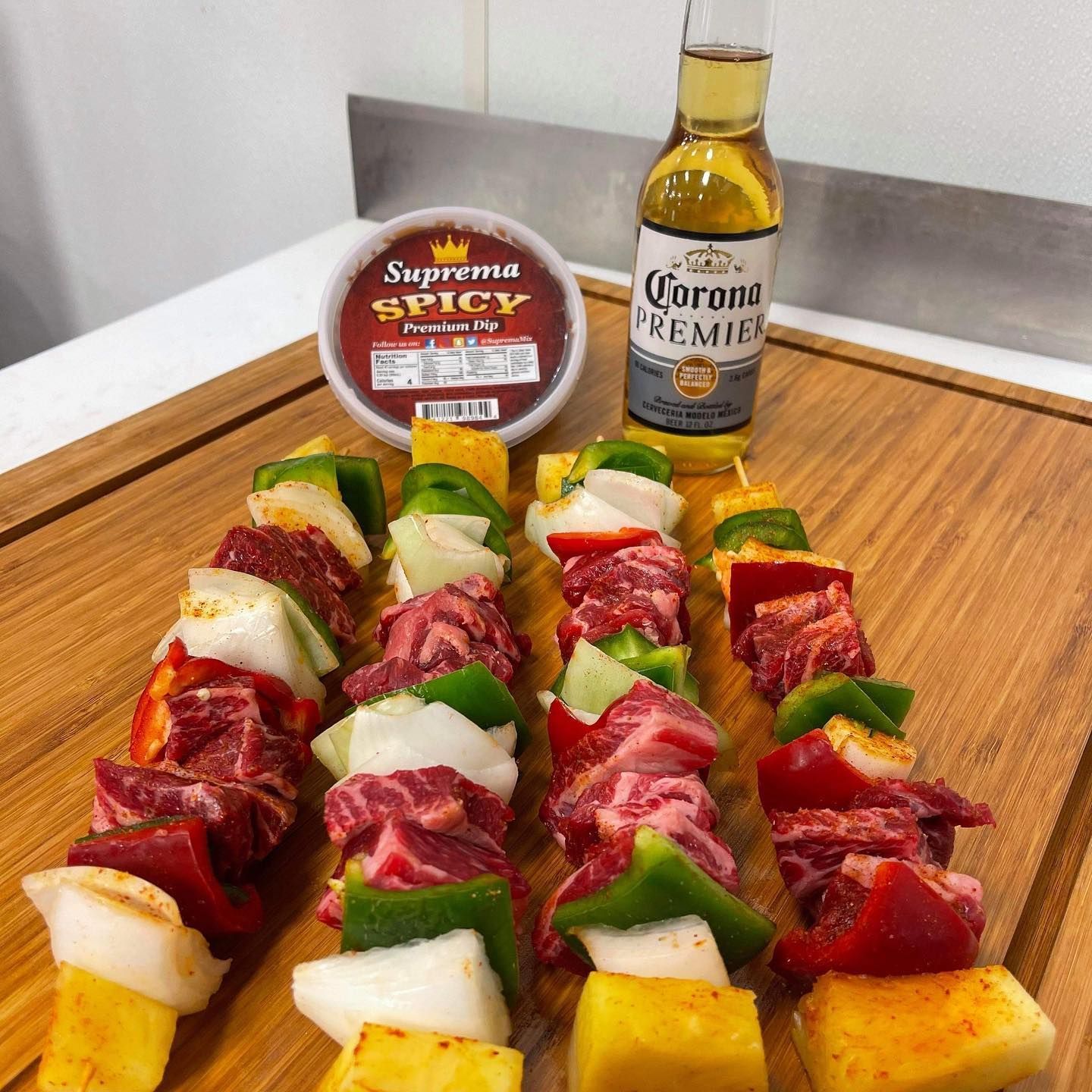 Assorted meat-and-fruit skewers on a wooden board with spicy dip and a Corona Premier bottle nearby
