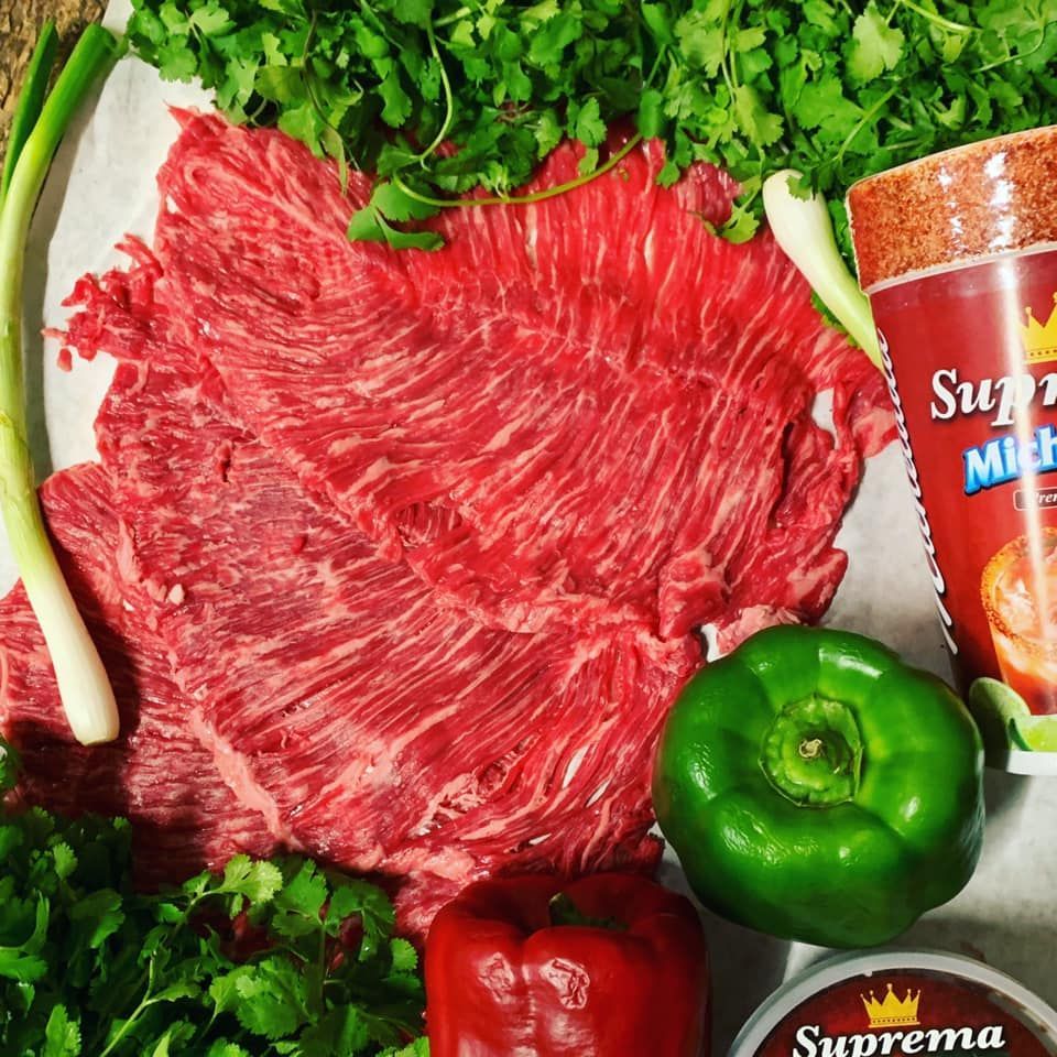 Raw beef steak surrounded by green herbs, peppers, and seasoning jars on a countertop