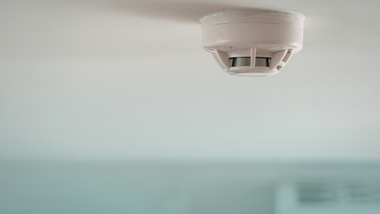 Ceiling-mounted smoke detector on a white ceiling with a blurred room background