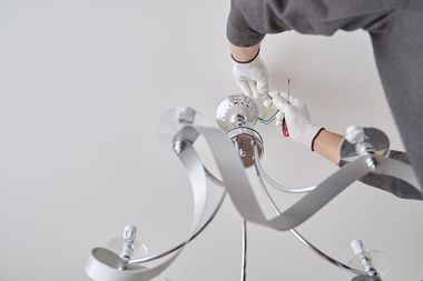 Person in white gloves installing a silver chandelier on a white ceiling.