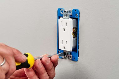 Hand tightening a wall outlet into a blue electrical box with a screwdriver