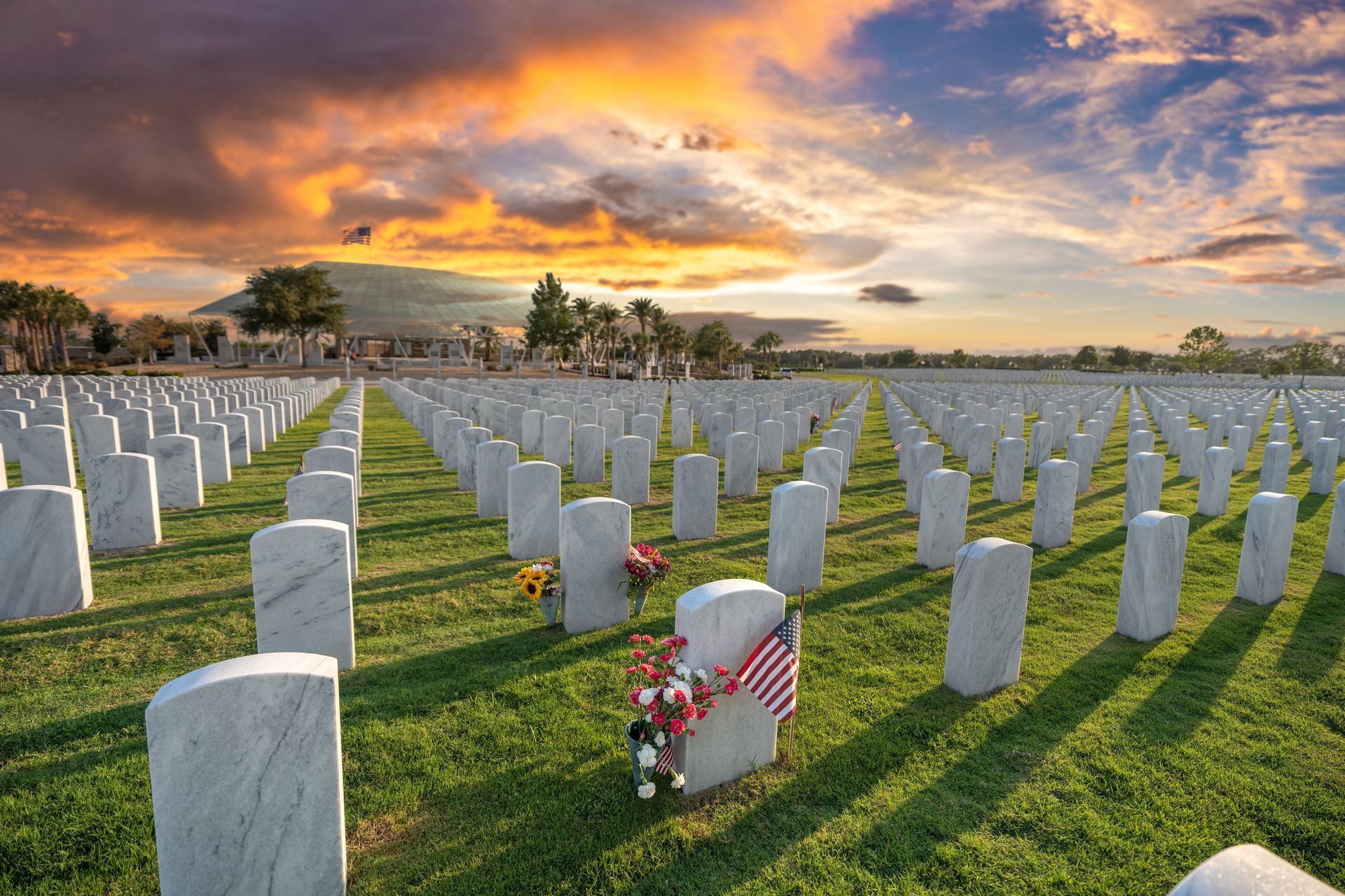 Rows of white gravestones in a cemetery at sunset, with one red-and-white wreath in the foreground.