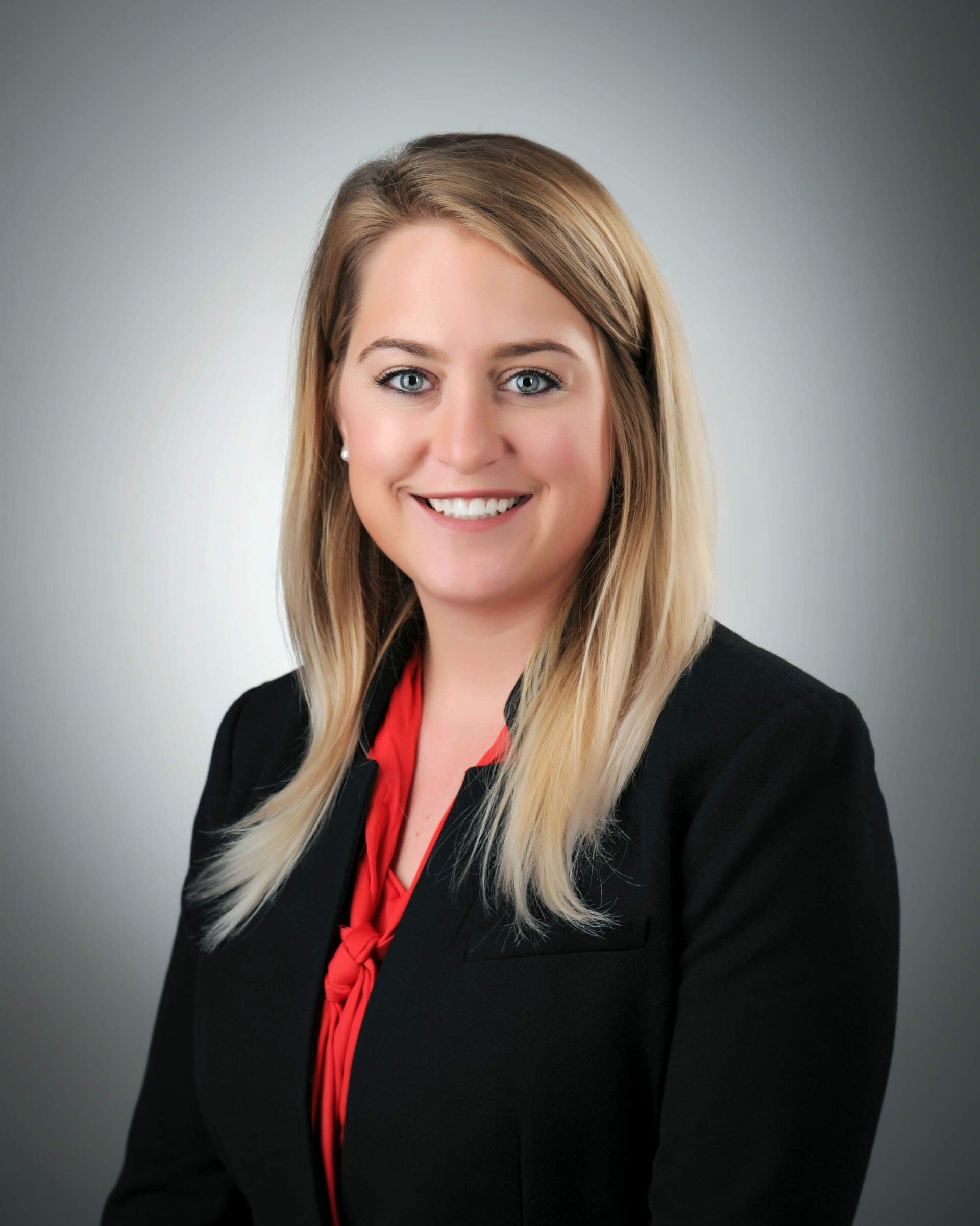 Woman with blonde hair wearing a black blazer and red blouse, smiling, studio setting.