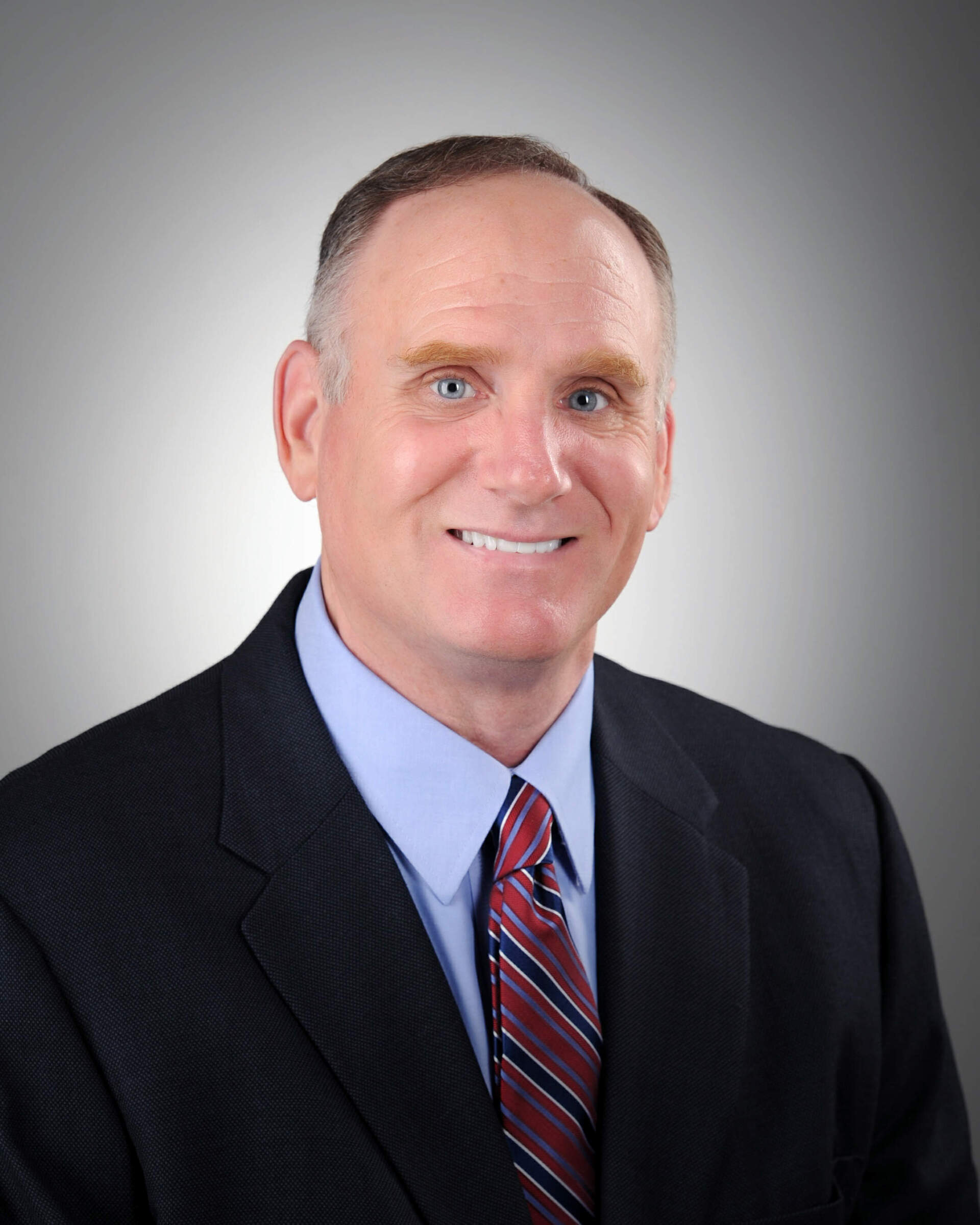 Man in suit smiles at the camera; blue shirt, red striped tie, gray gradient background.