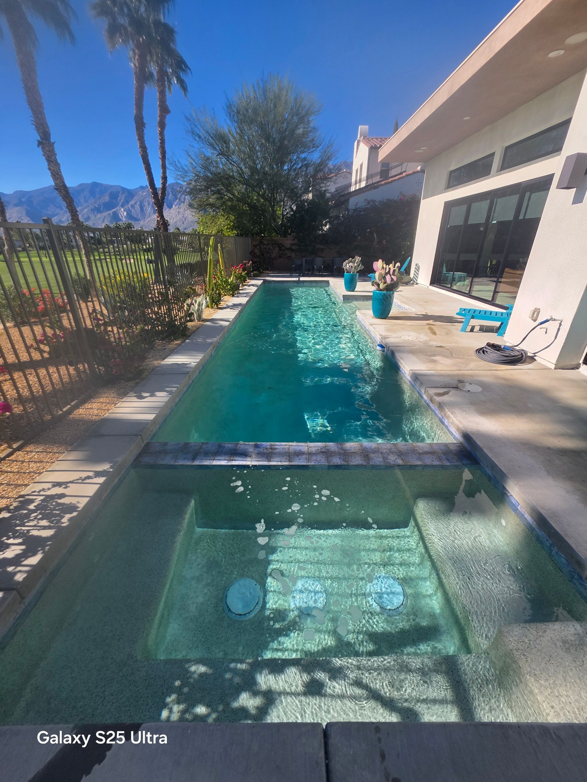 Narrow, turquoise pool with steps, next to a house with a concrete patio. Palm trees and mountains in background.
