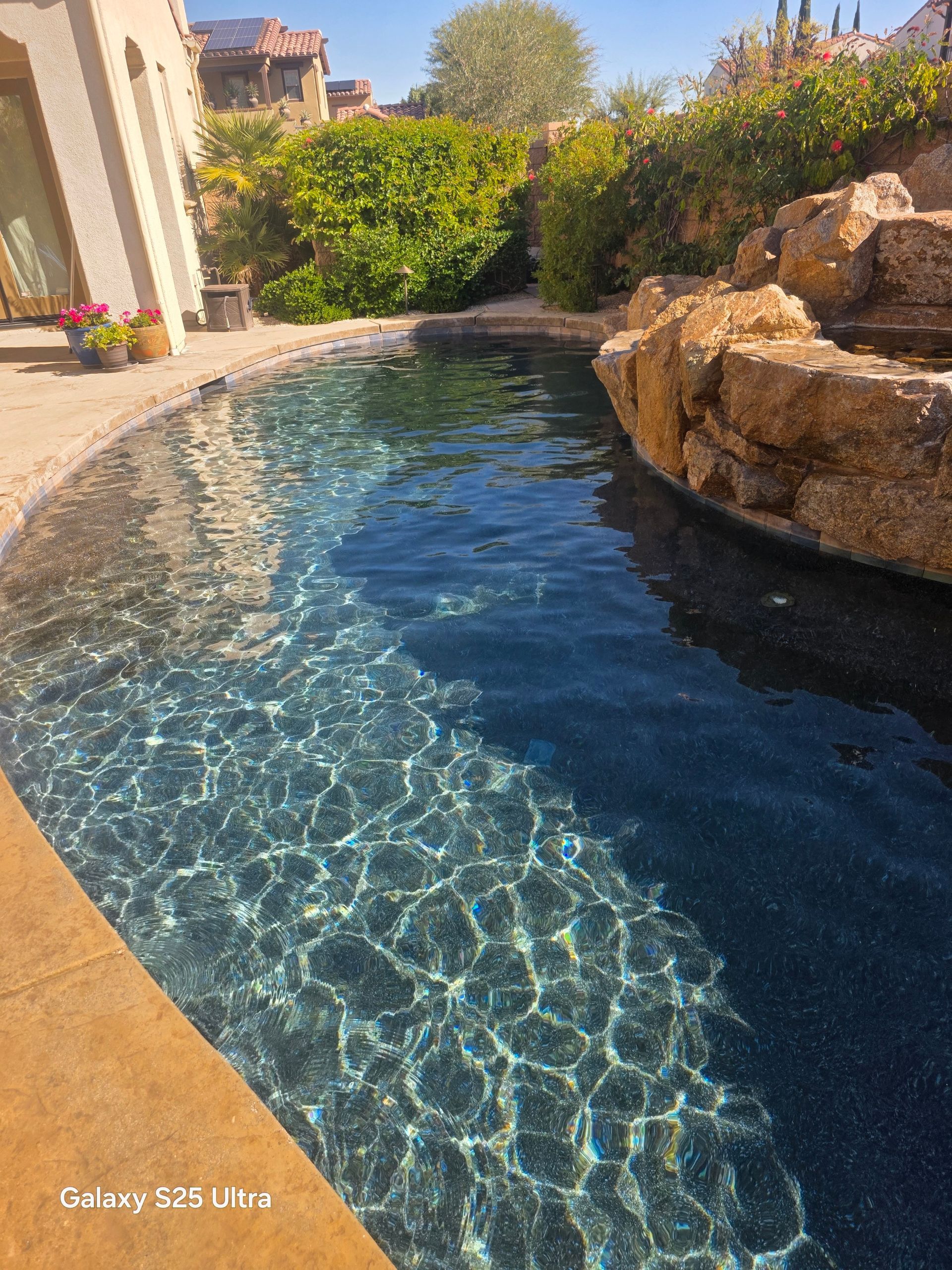 Pool with clear water reflecting sunlight, edged by tan stone and large rocks.
