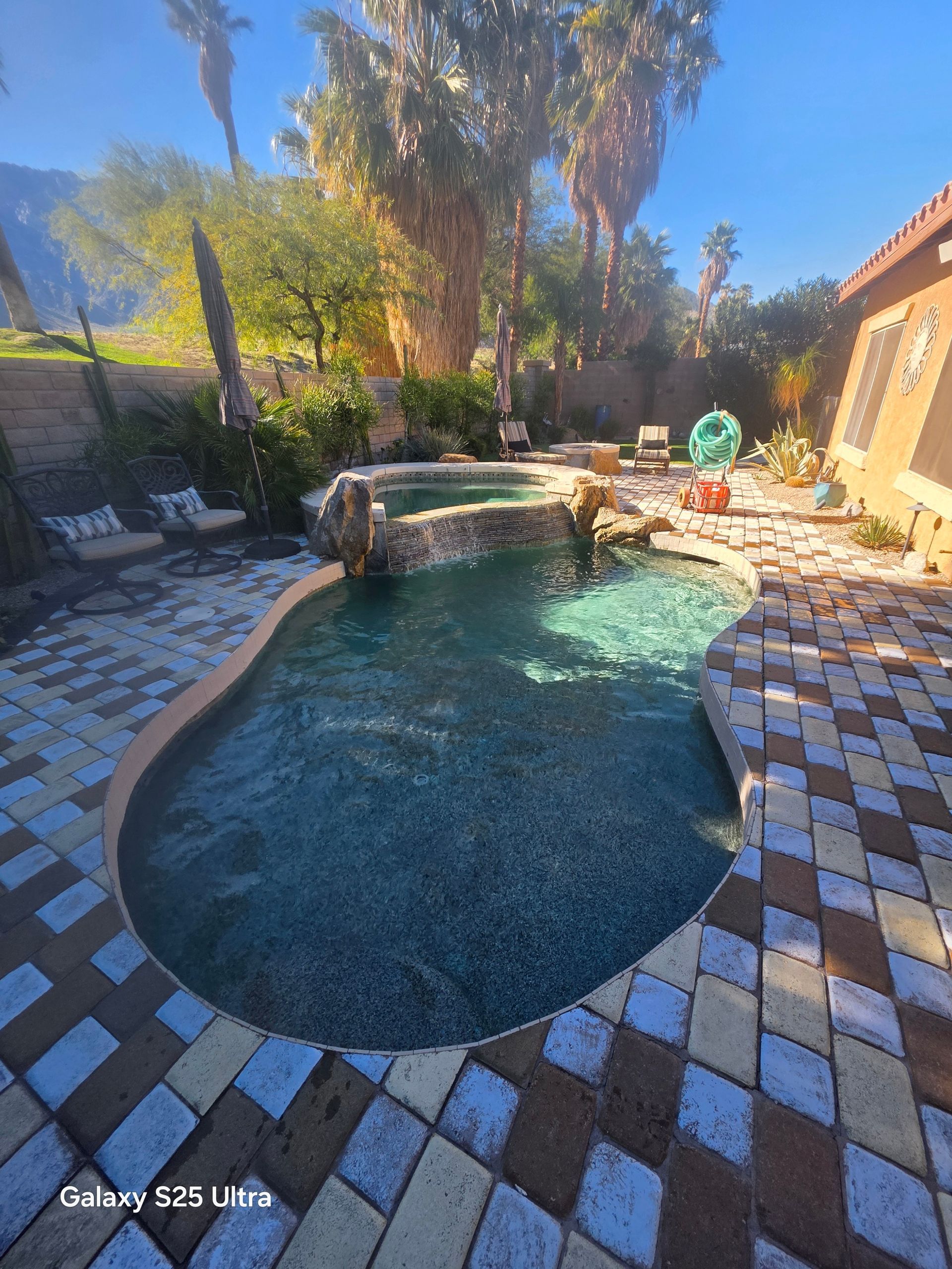 Pool with waterfall feature and surrounding stone patio in a sunny backyard with trees.