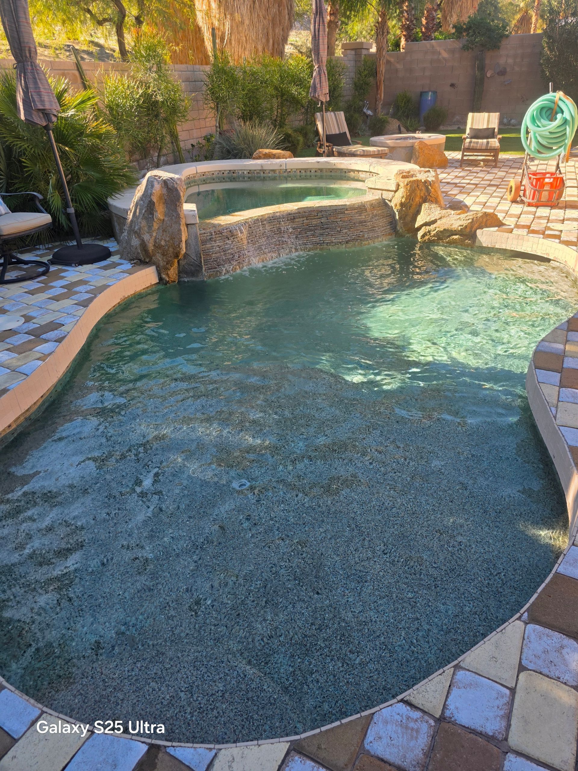 Pool with cascading waterfall, surrounded by stone patio, in a sunny outdoor setting.