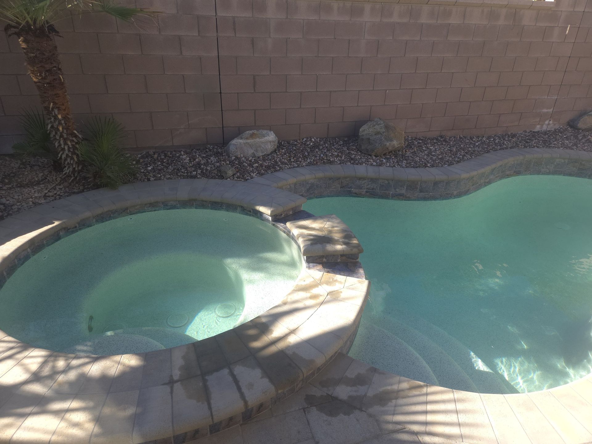 Pool with attached spa, surrounded by stone and a brick wall. Water is turquoise and clear.