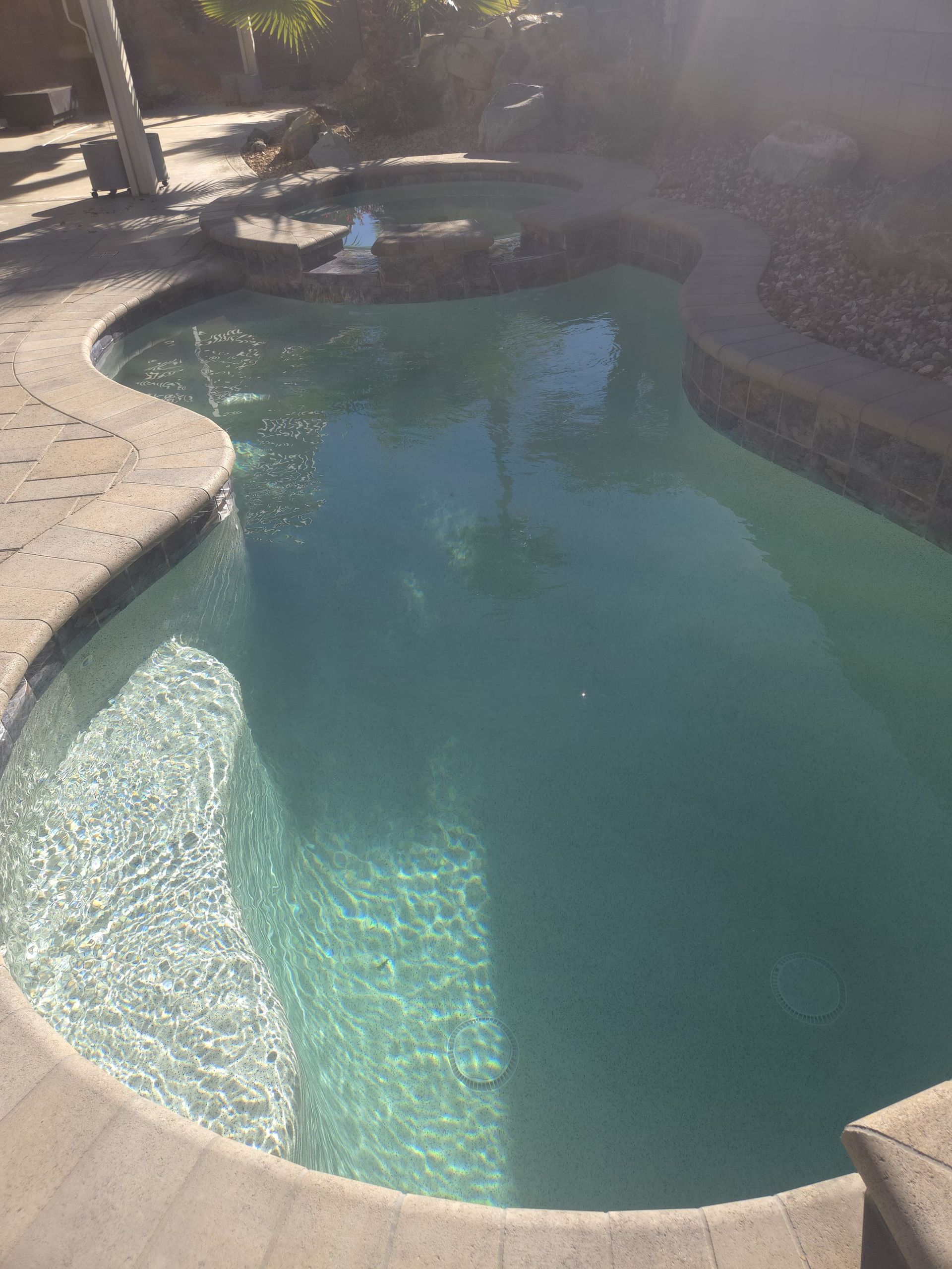A kidney-shaped swimming pool with clear turquoise water and a small hot tub. Concrete deck surrounds.