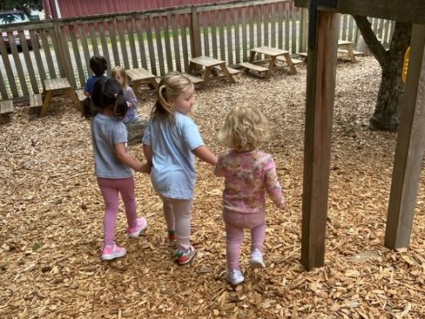 montessori children walk hand in hand on a woodchip-covered playground near picnic tables and a red 