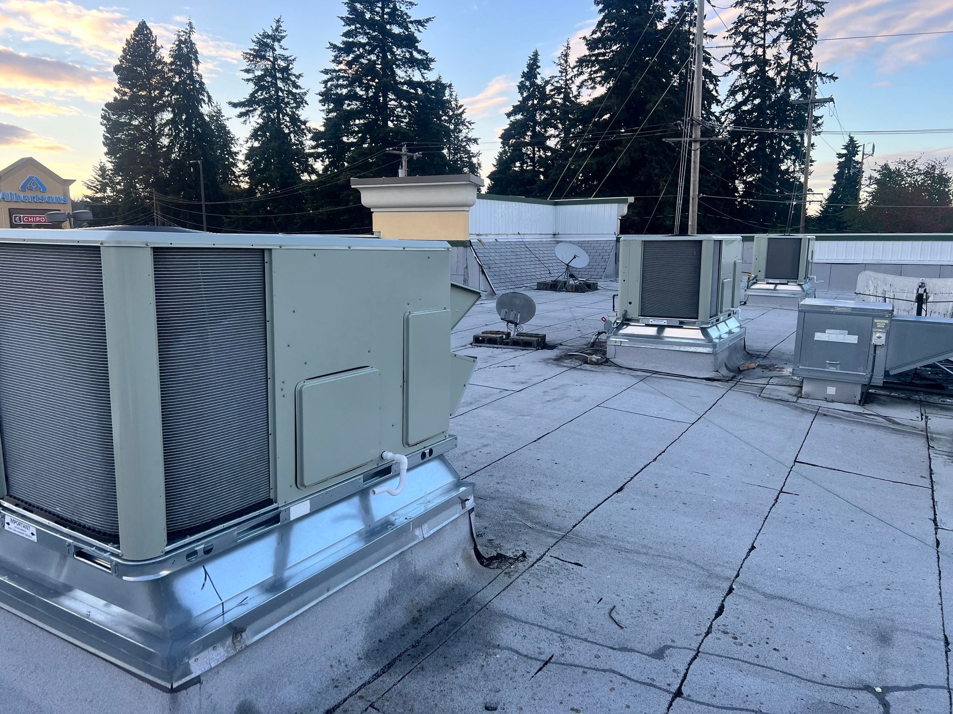 Rooftop HVAC units on a flat commercial roof at dusk, with trees and sky in the background
