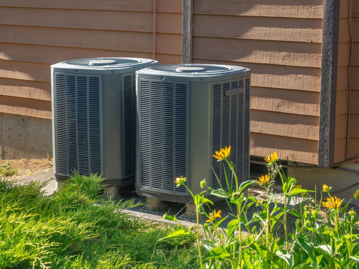 Two air conditioners are sitting next to each other on the side of a house.