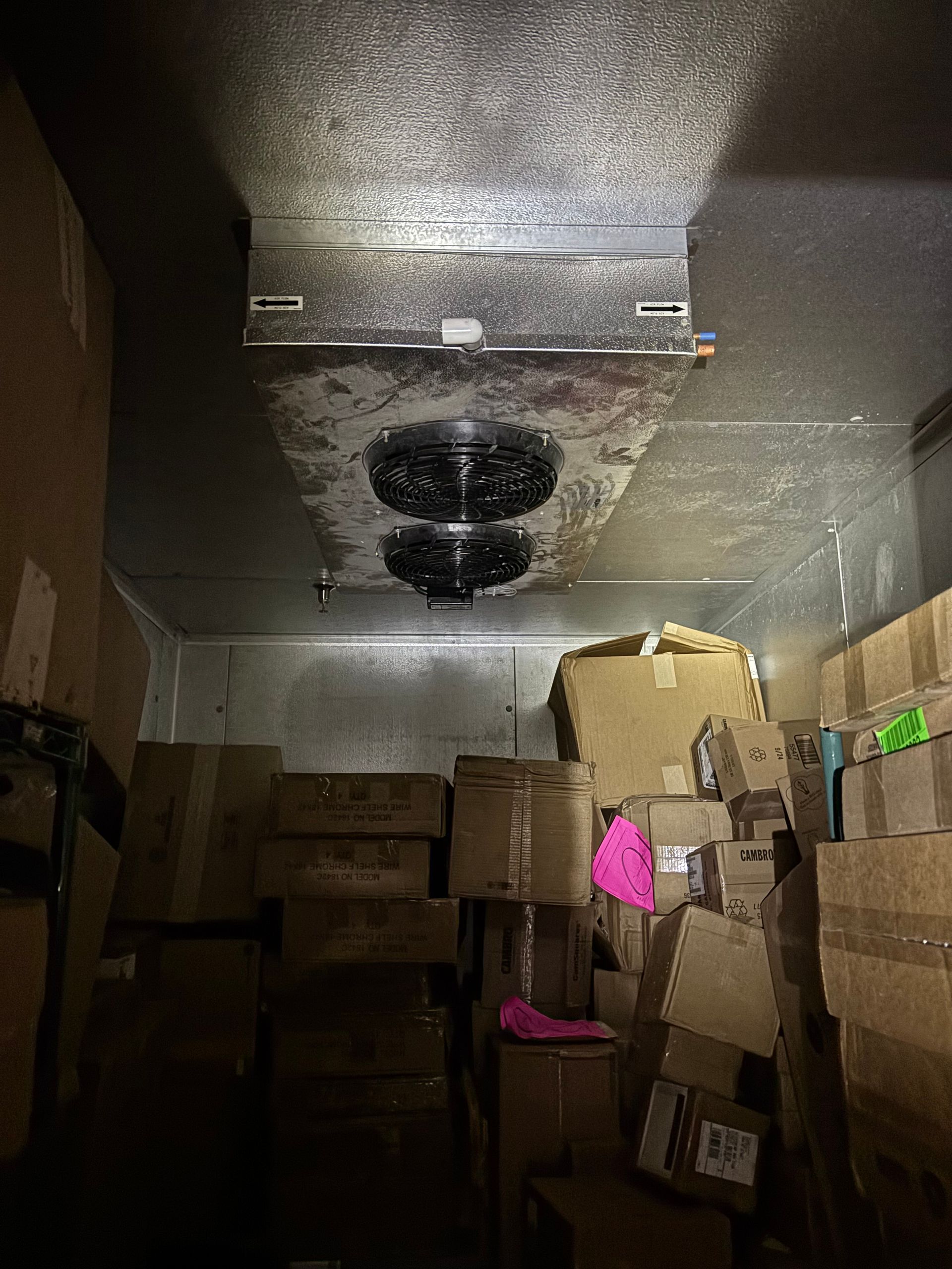 Dim storage room with stacked cardboard boxes and two black ceiling fans on a reflective metal ceiling.