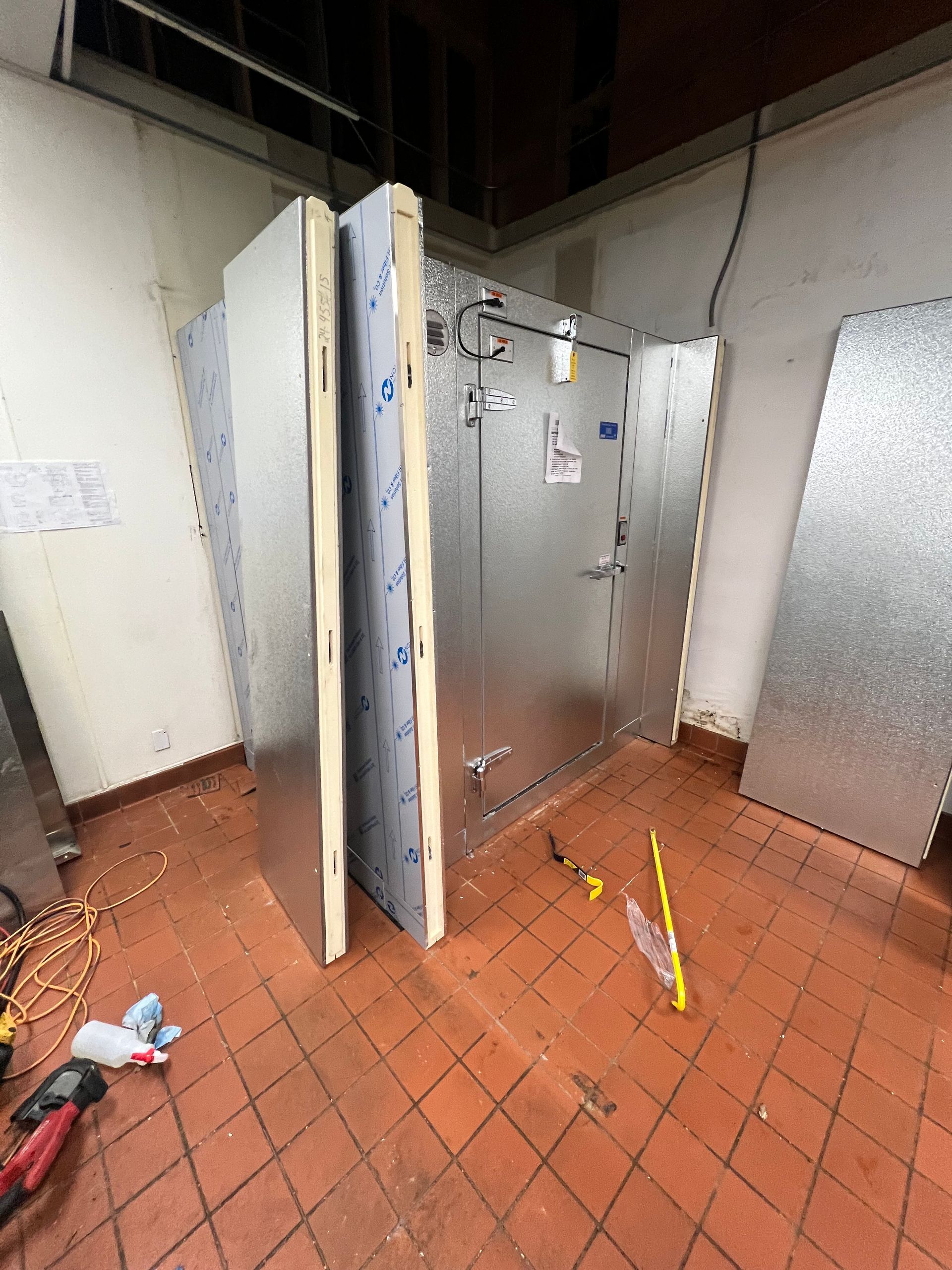 Industrial room with a metal door ajar, large panels, and tools on a reddish tiled floor.