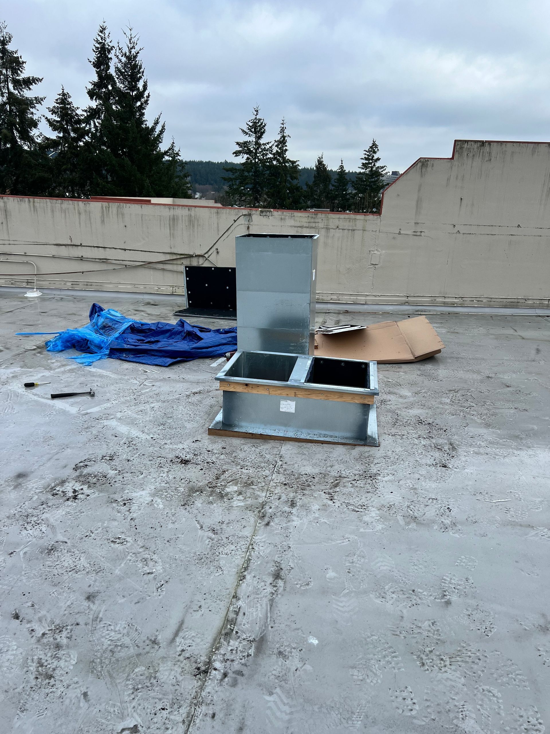 Damaged appliance parts on a gravel lot beside a low concrete wall, with blue tarp and trees in background