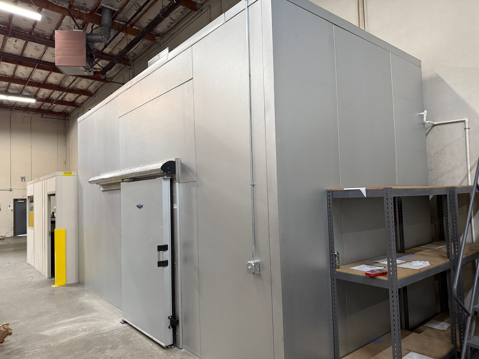 Industrial walk-in cooler with gray metal walls, a white door, and a storage shelf in a warehouse aisle