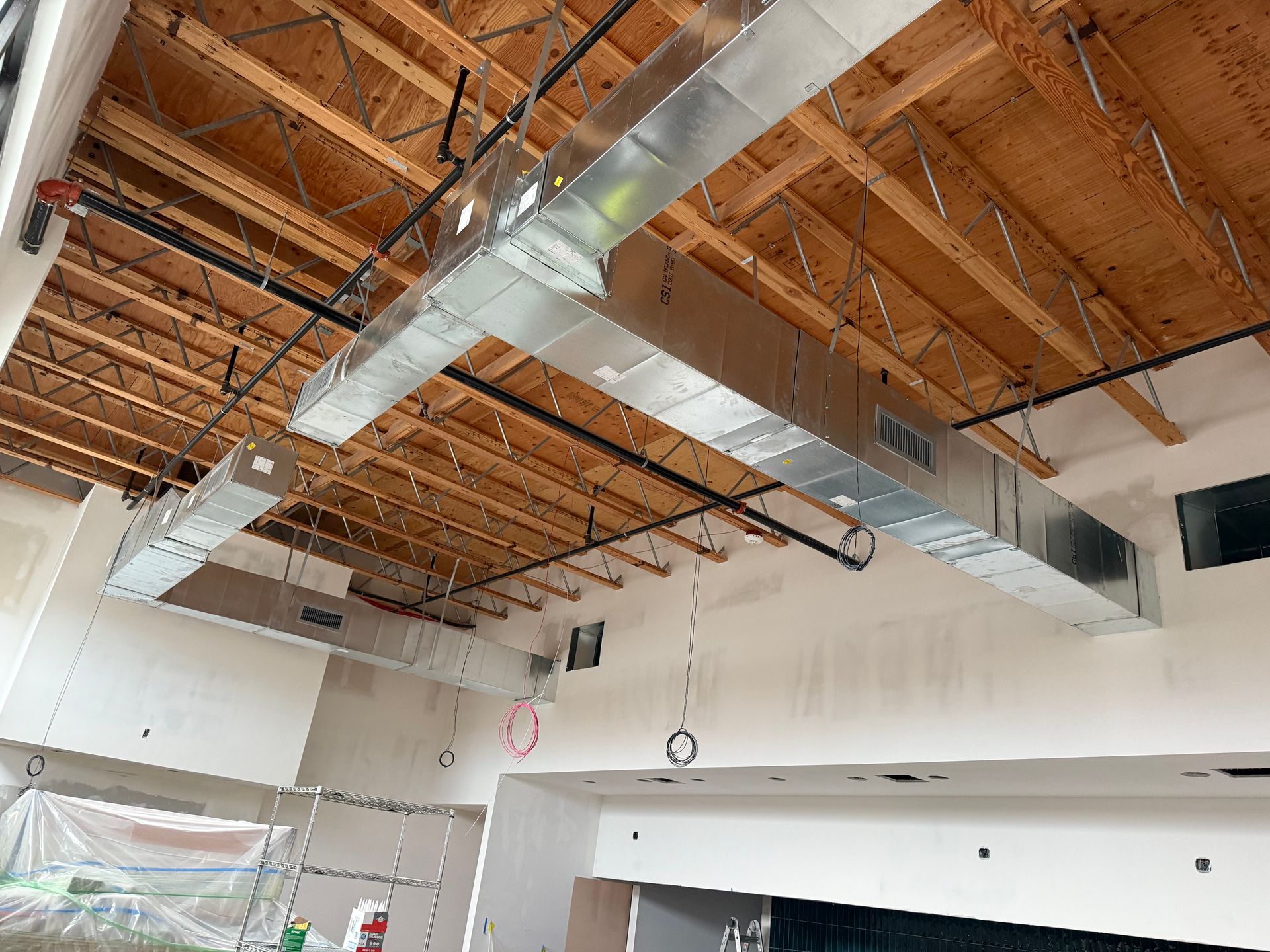 Interior under construction with exposed wooden trusses, silver ductwork, and unfinished white walls.