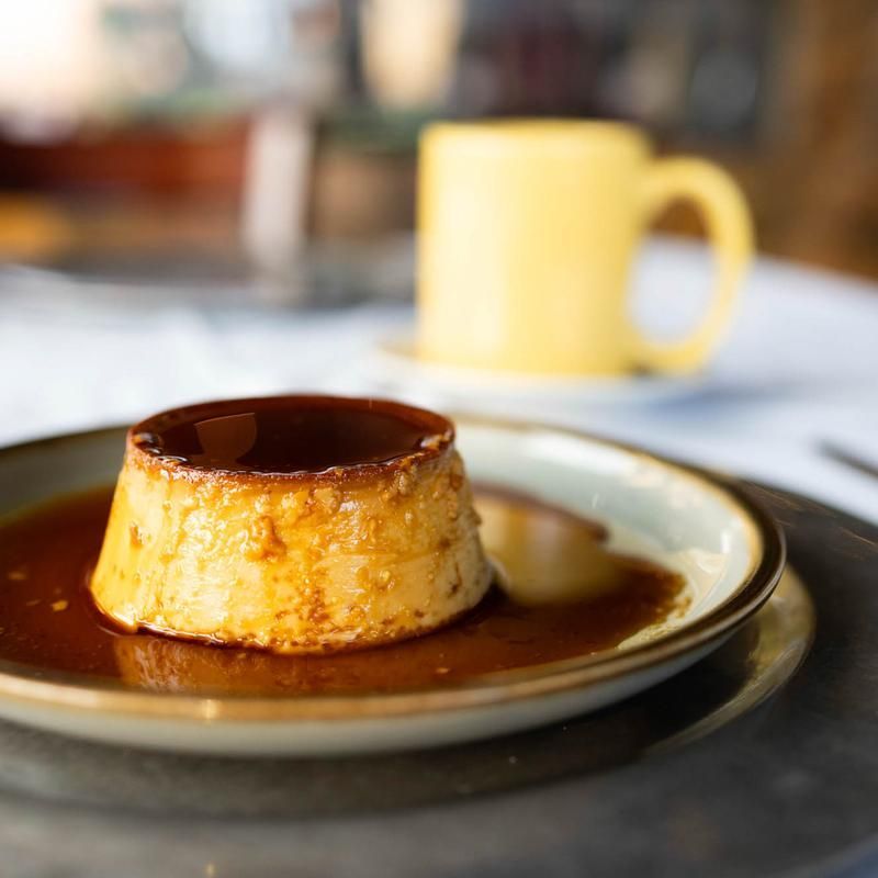 Caramel flan on a plate with sauce, with a yellow mug blurred in the background.