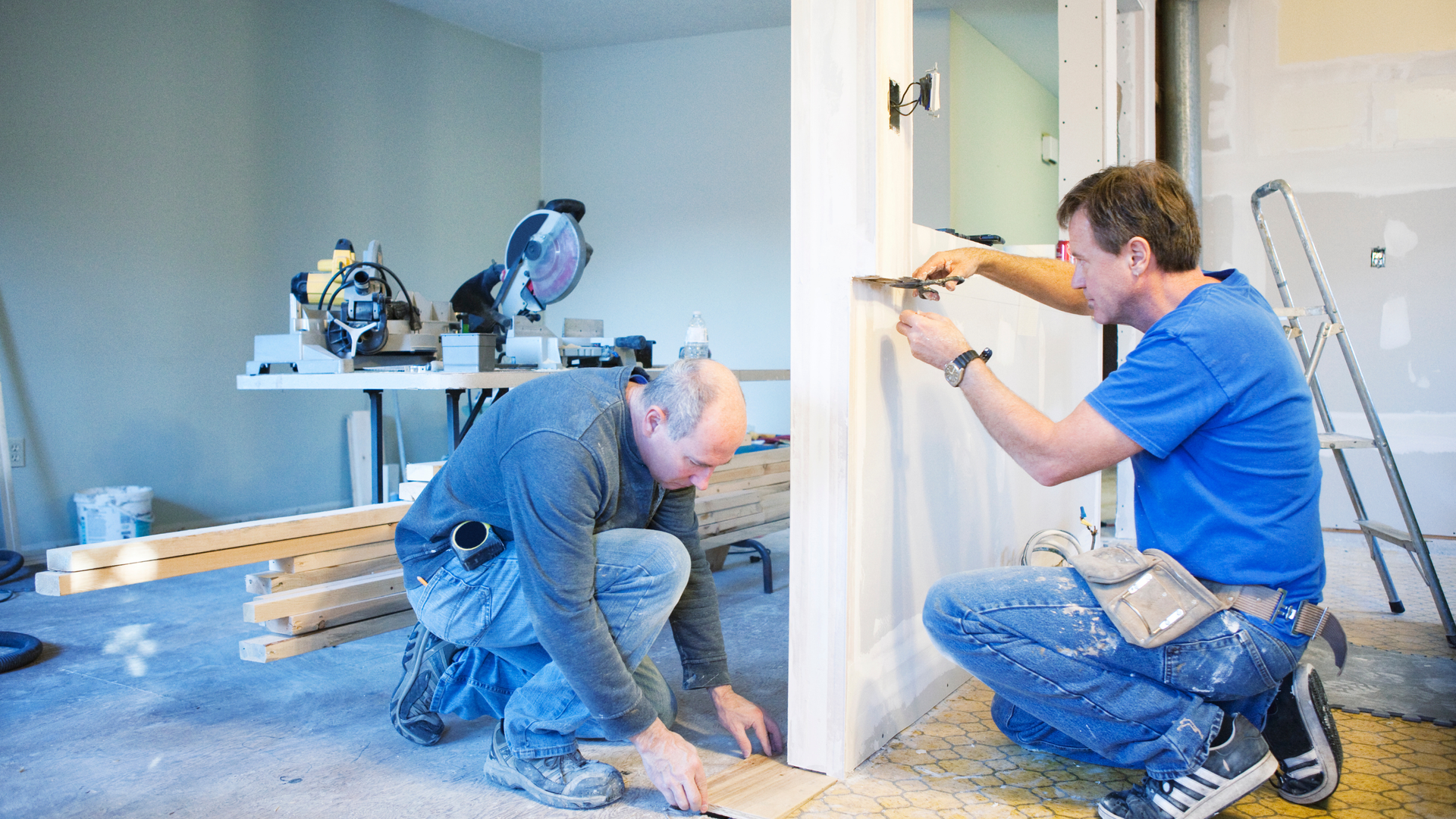 Two workers installing white trim in a bright room, kneeling with power tools and measuring a wall.