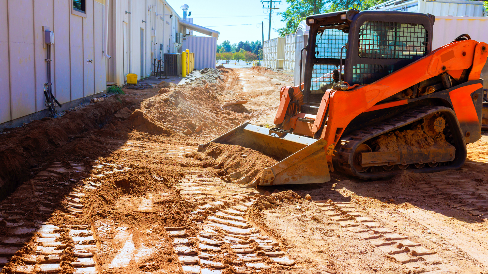 Orange skid steer working on dirt path beside a building on a construction site